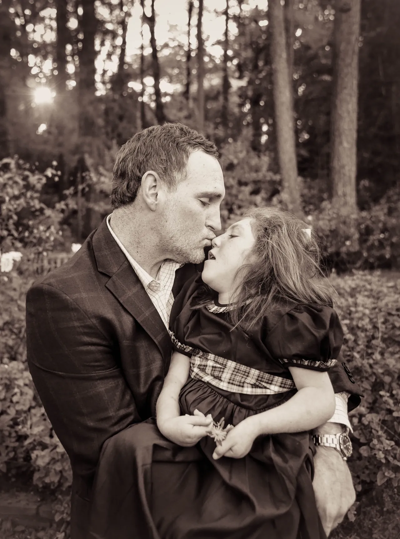 Sepia portrait of a man holding and kissing a young girl in a forest garden at sunset.