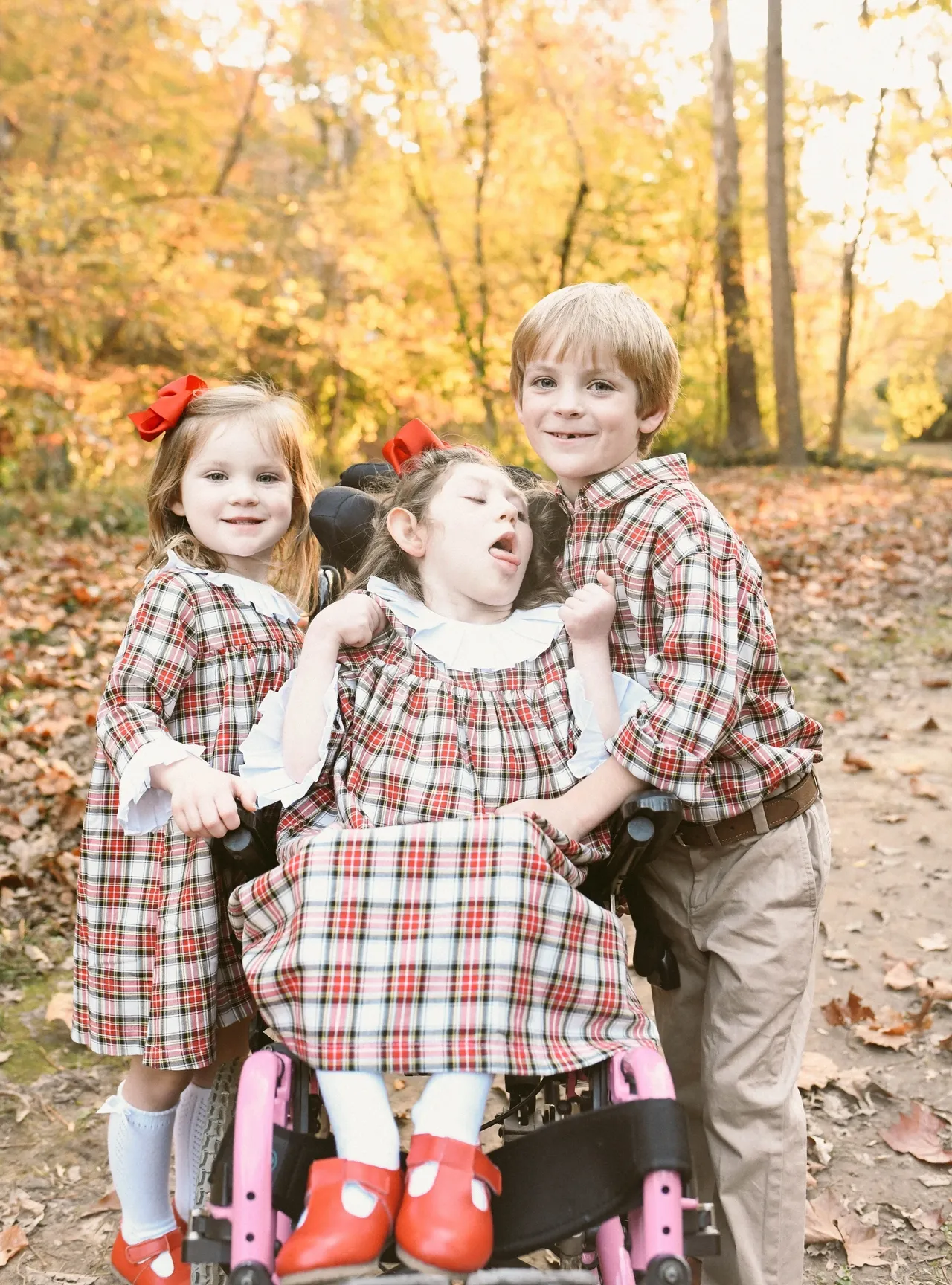 Three children in matching plaid outfits posing on a forest path with golden fall leaves