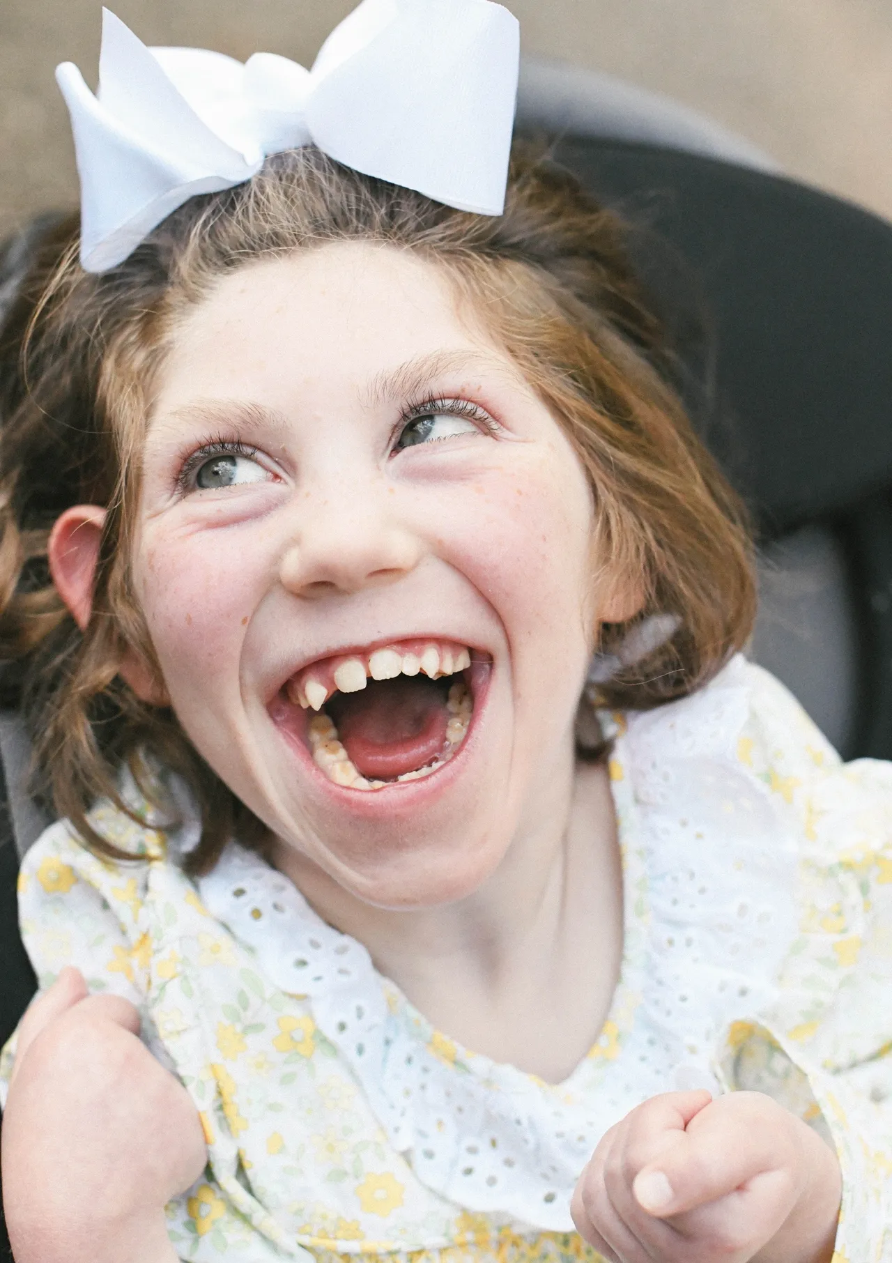 Smiling child with white hair bow laughing in a floral dress with eyelet collar, close-up portrait.