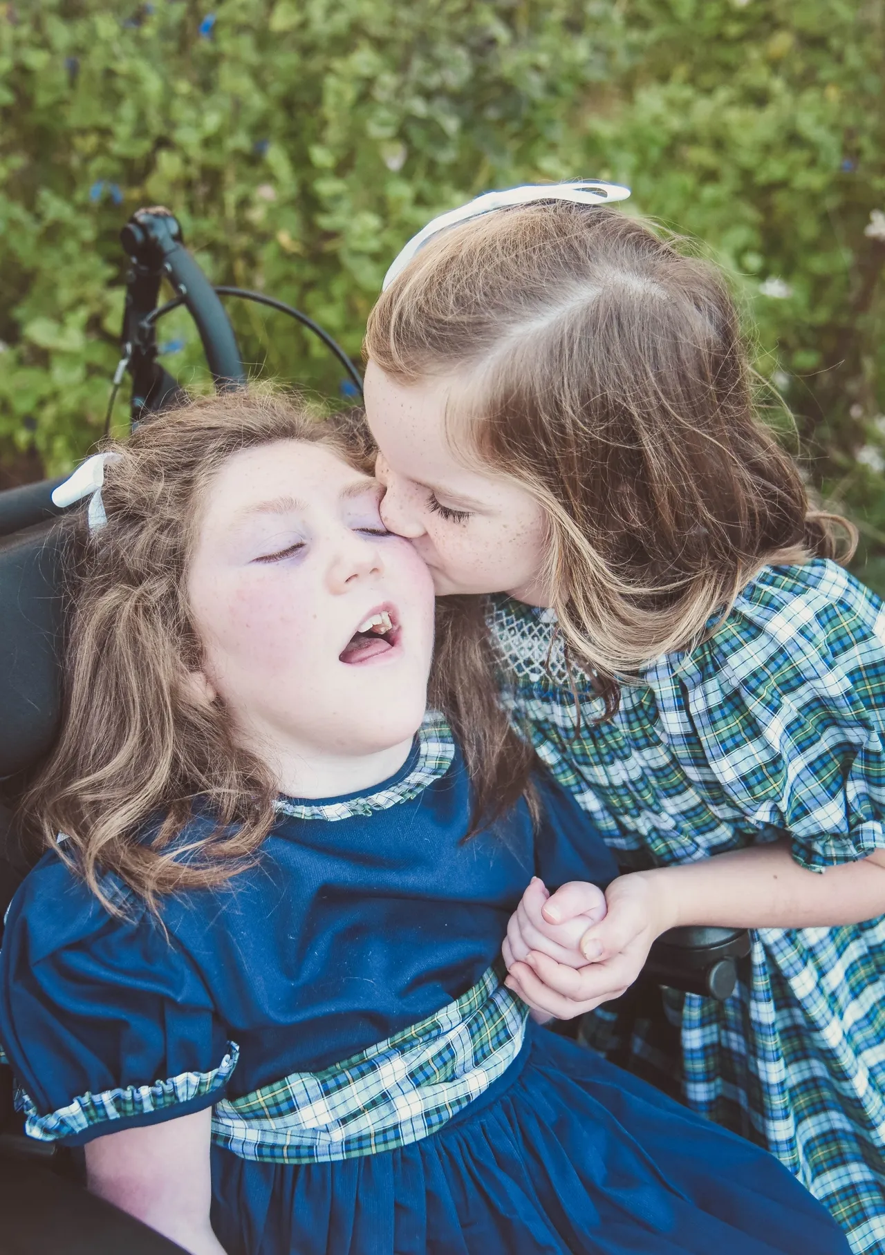 Two children in matching blue plaid dresses; one kisses the other's cheek while holding hands outdoors