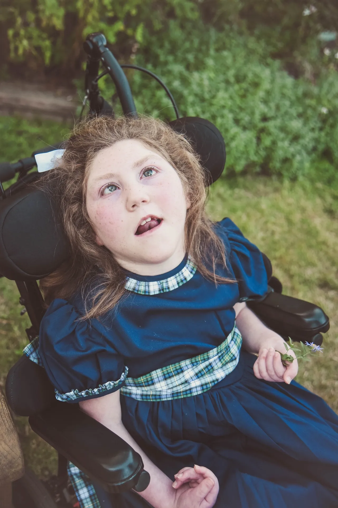 A child in a wheelchair wearing a blue plaid-trim dress holds a small purple flower outdoors.