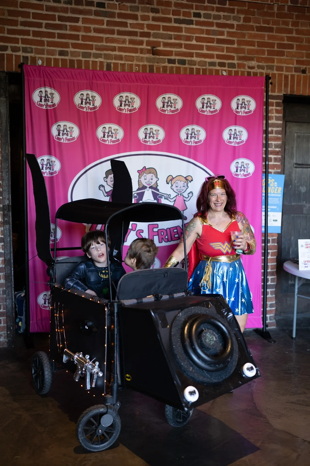 Woman in Wonder Woman costume with two kids in Batmobile stroller at event with pink backdrop.