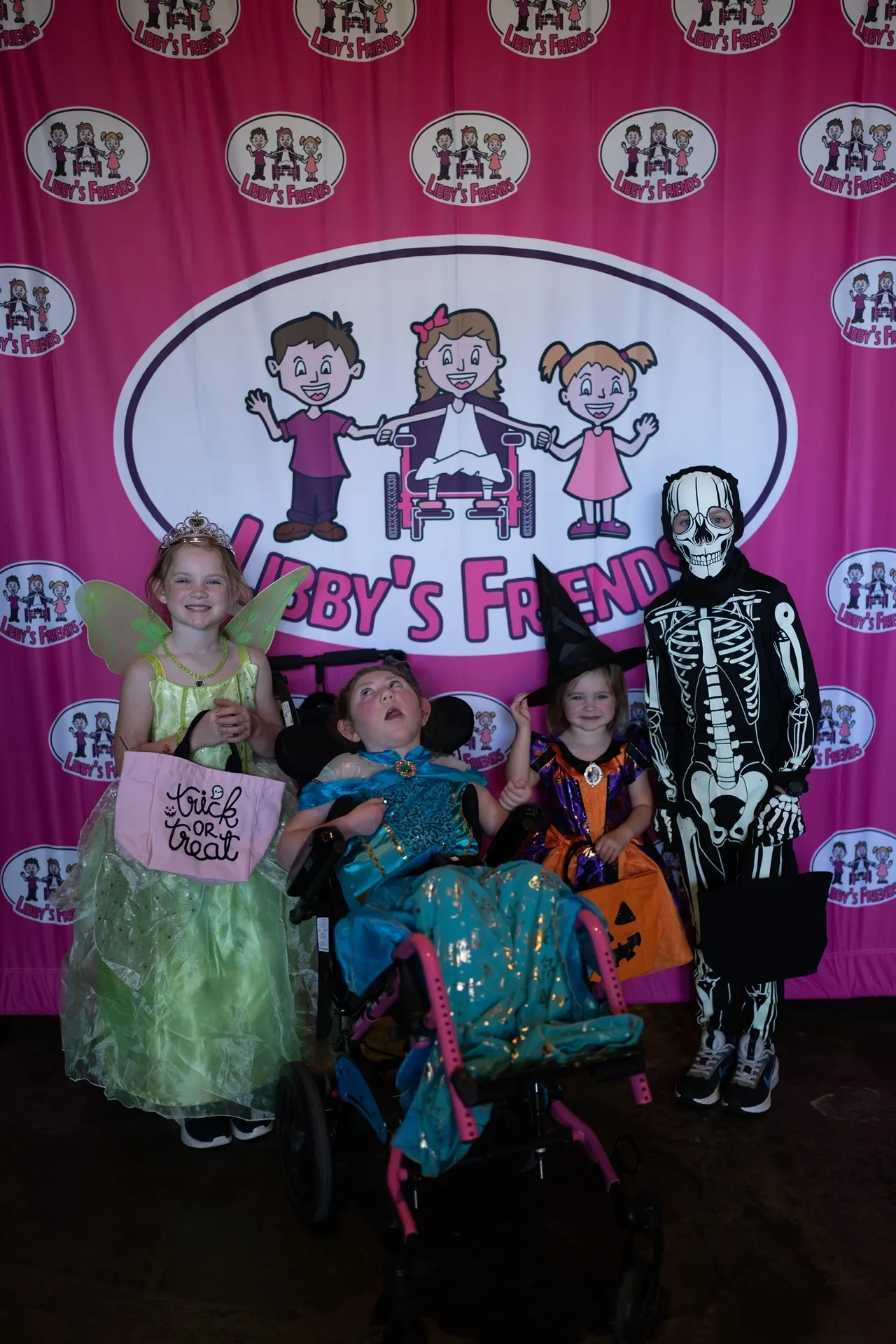 Four children in fairy, princess, witch, and skeleton costumes by a pink Libby’s Friends backdrop, one in a wheelchair.