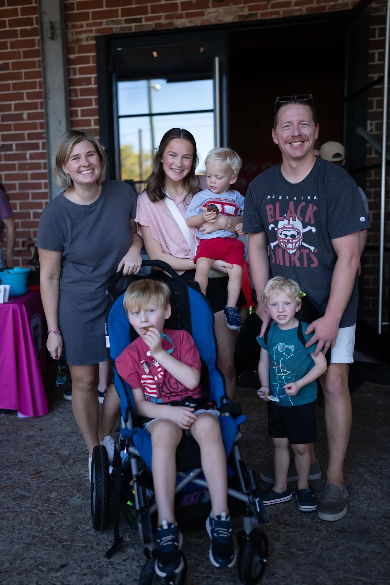 Smiling family with parents, a teen, and three young kids by a brick building, one child in a stroller at a community event