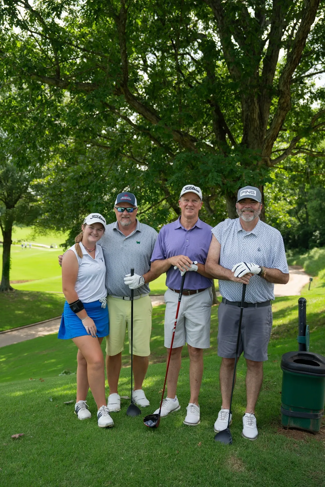Four golfers posing with drivers on a sunny, green course under trees, smiling before teeing off.