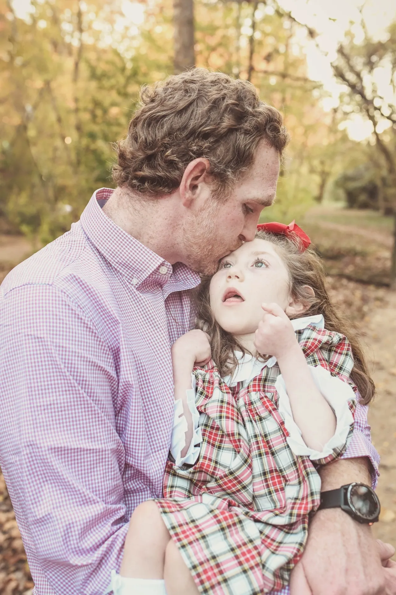 A man kissing a young girl’s forehead while holding her, plaid dress and red bow, in an autumn park.