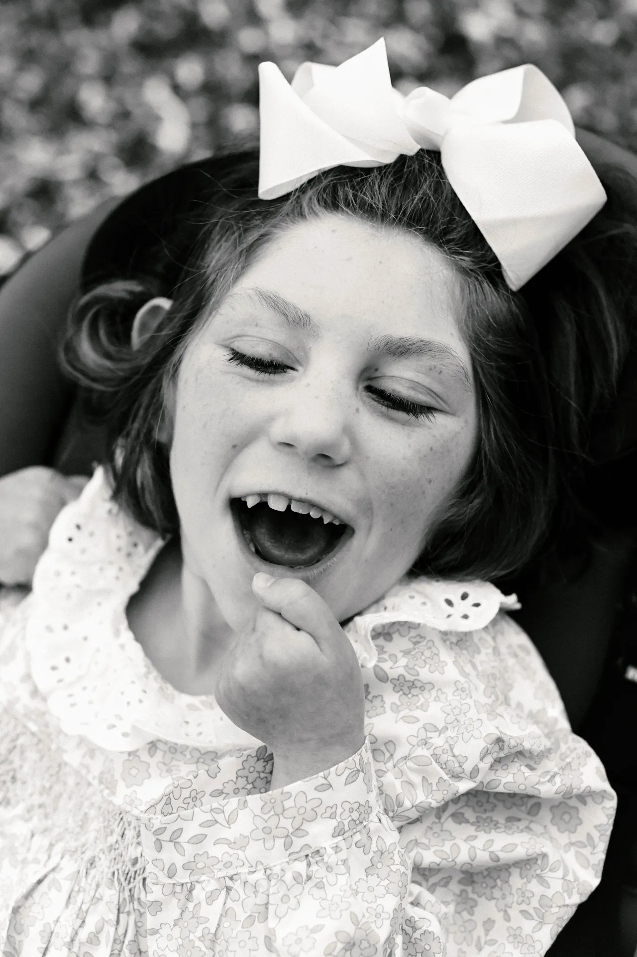 Black-and-white portrait of a smiling child with a big hair bow, floral dress, and open mouth, laughing outdoors.