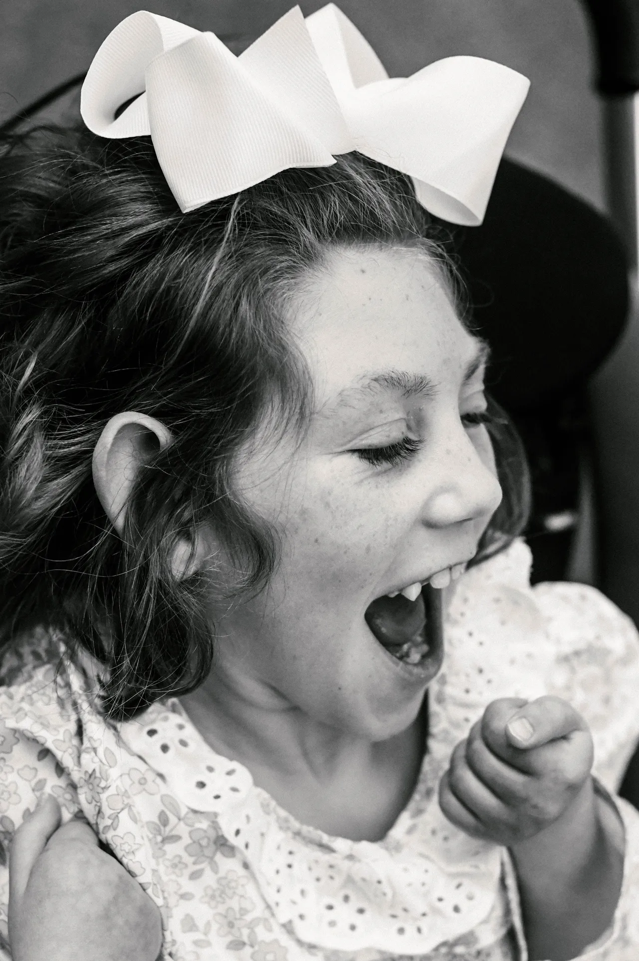 Black-and-white close-up of a joyful child laughing, wearing a large white hair bow and floral dress.