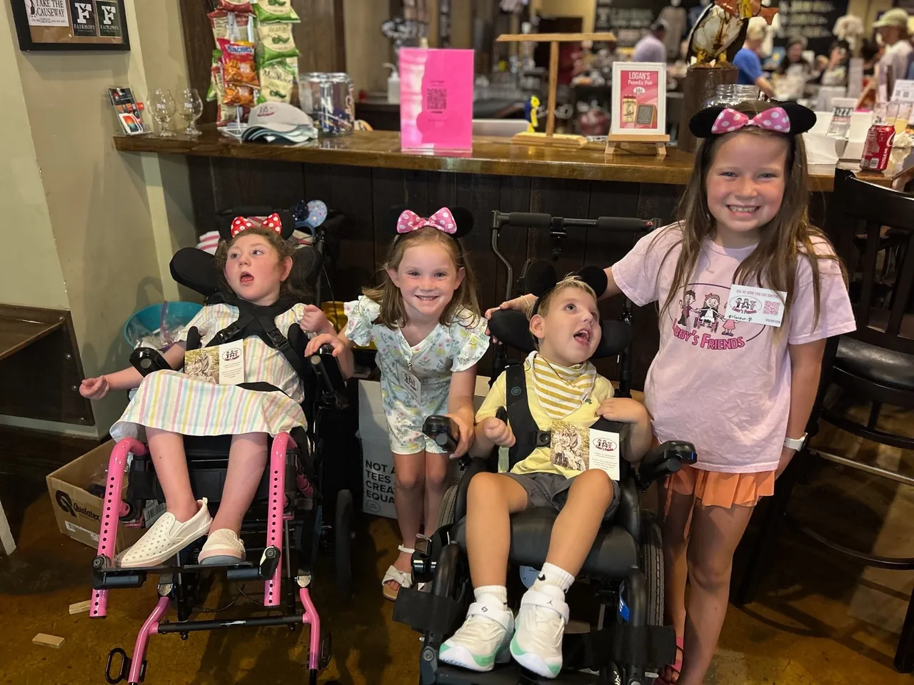 Four children at a restaurant, two in wheelchairs, wearing mouse ear headbands, smiling by the bar.