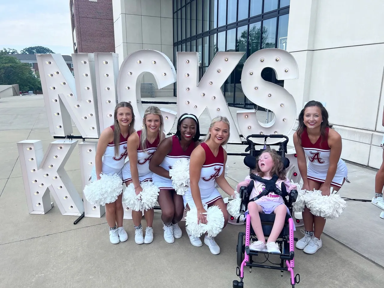University of Alabama cheerleaders pose with a child by the NICKS KIDS marquee sign, holding white pom-poms.