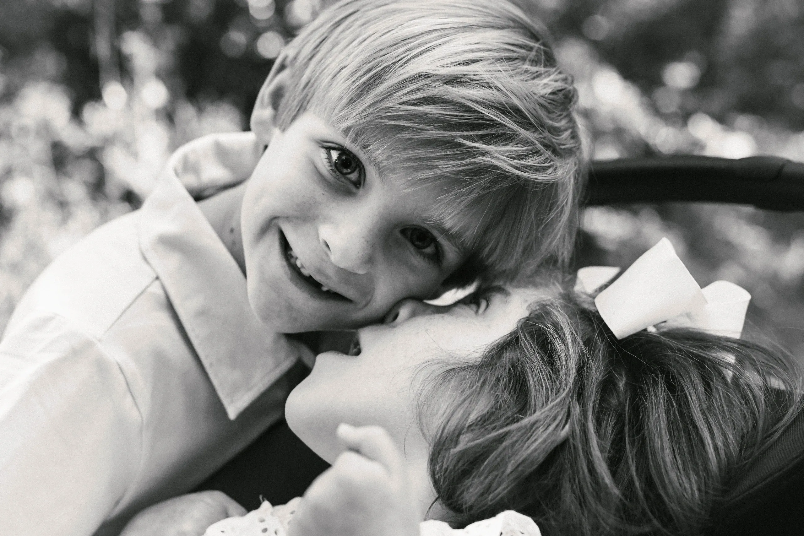 Black-and-white portrait of two children cuddling; a smiling boy leans over a girl with a bow outdoors