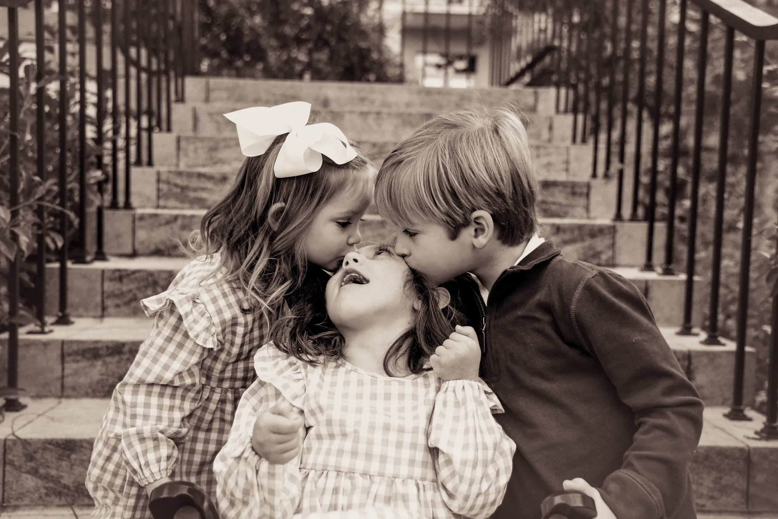 Siblings share kisses on sister's forehead on outdoor stone steps, heartwarming family portrait in sepia