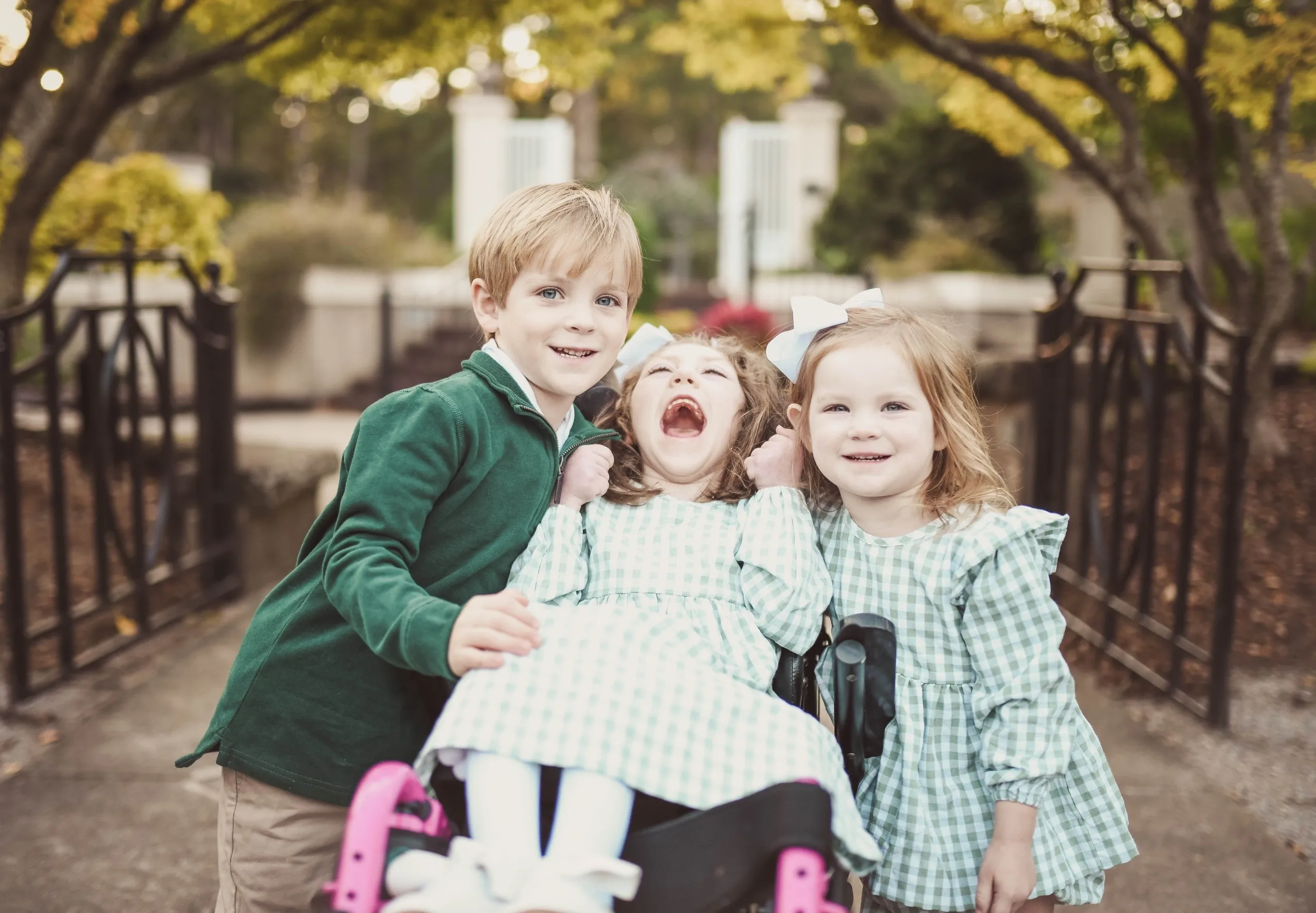 Three children smile outdoors; two girls in green gingham dresses, one in a wheelchair, with a boy in green by a garden gate.