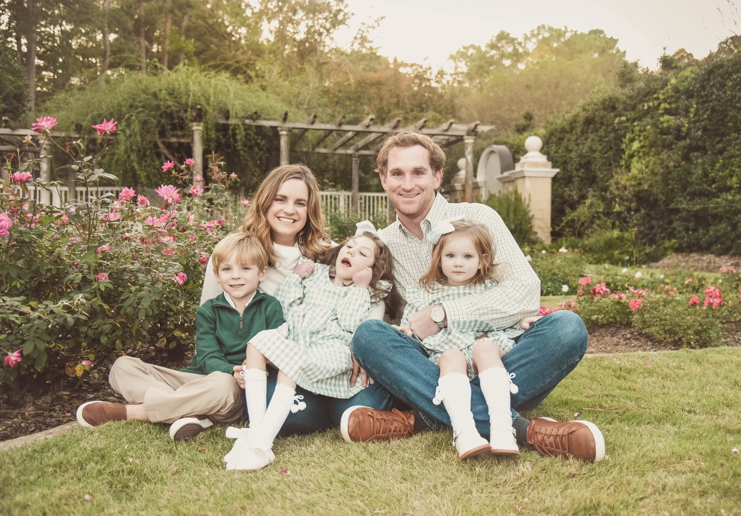 Family portrait: smiling parents with three kids, two in matching dresses, sitting in a rose garden at sunset.