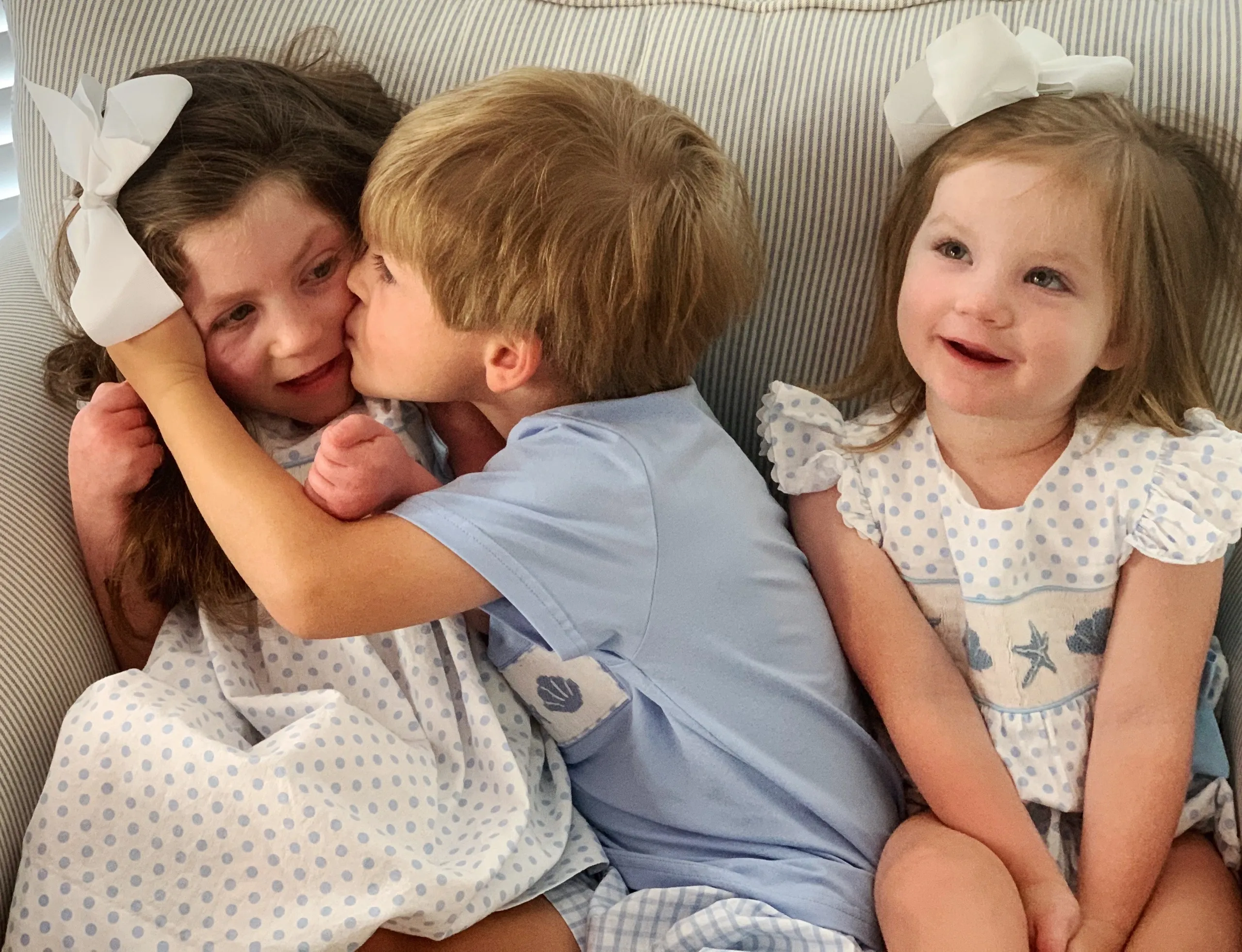 Boy kisses girl's cheek while another girl smiles; three children in blue and white outfits on a striped couch.