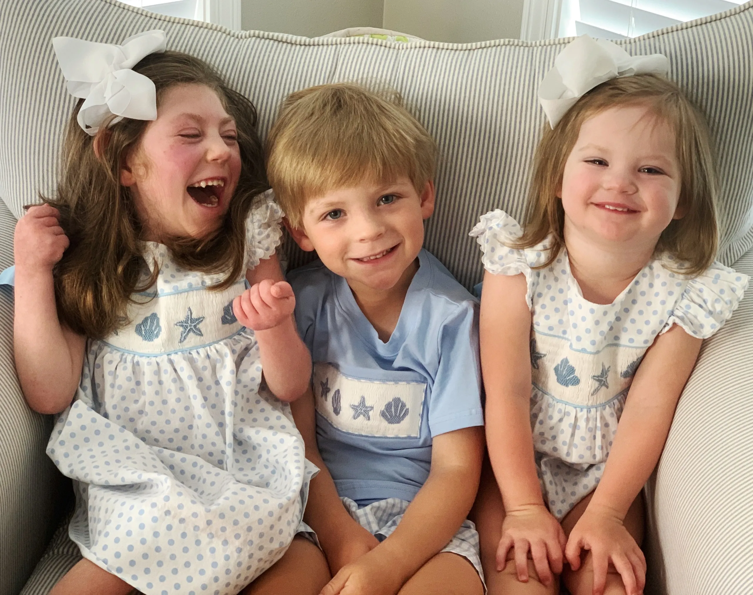 Three smiling siblings in matching blue and white seashell outfits on a striped couch