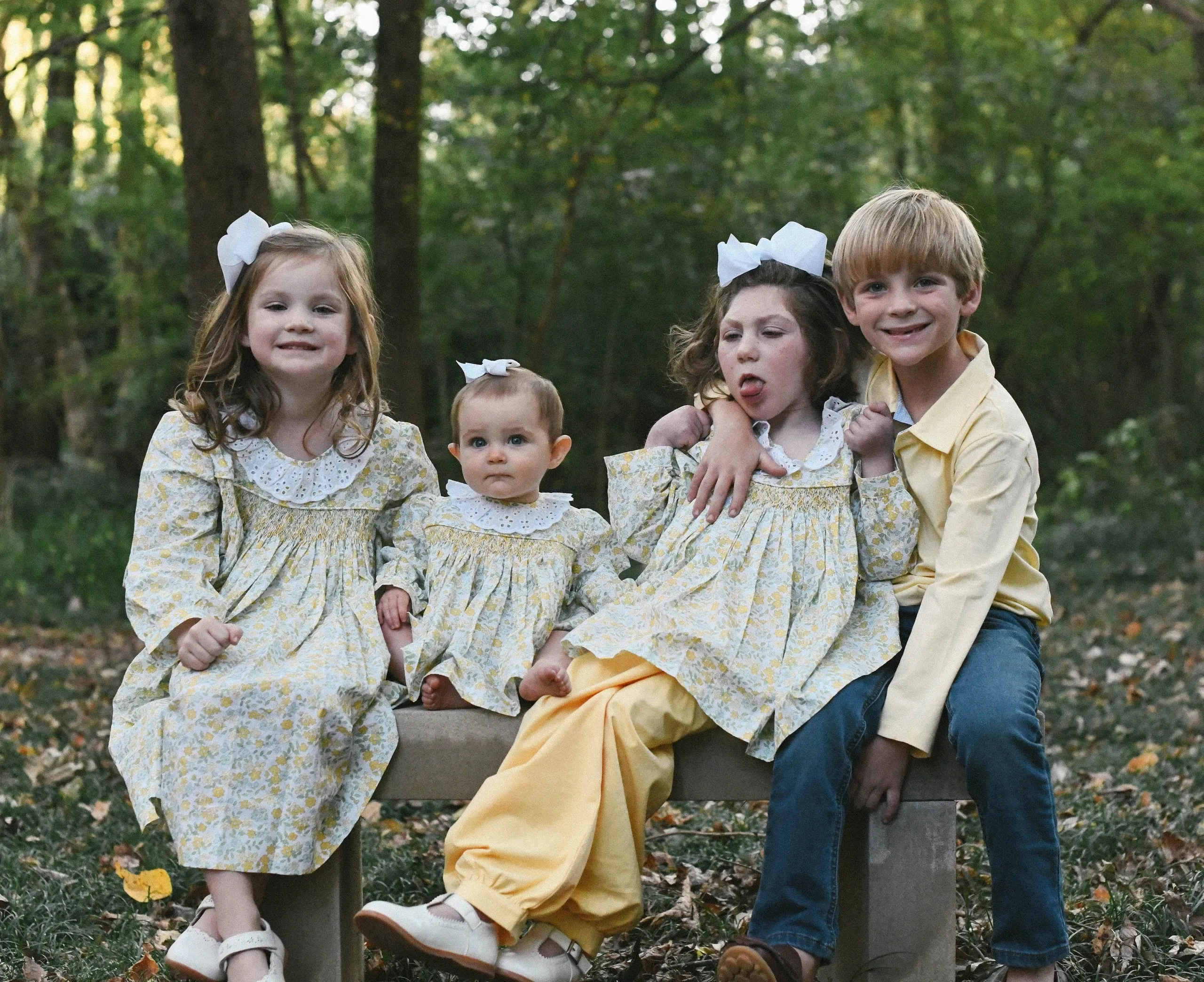 Four children on a bench, three girls in yellow floral dresses and a boy in yellow shirt, autumn woods backdrop