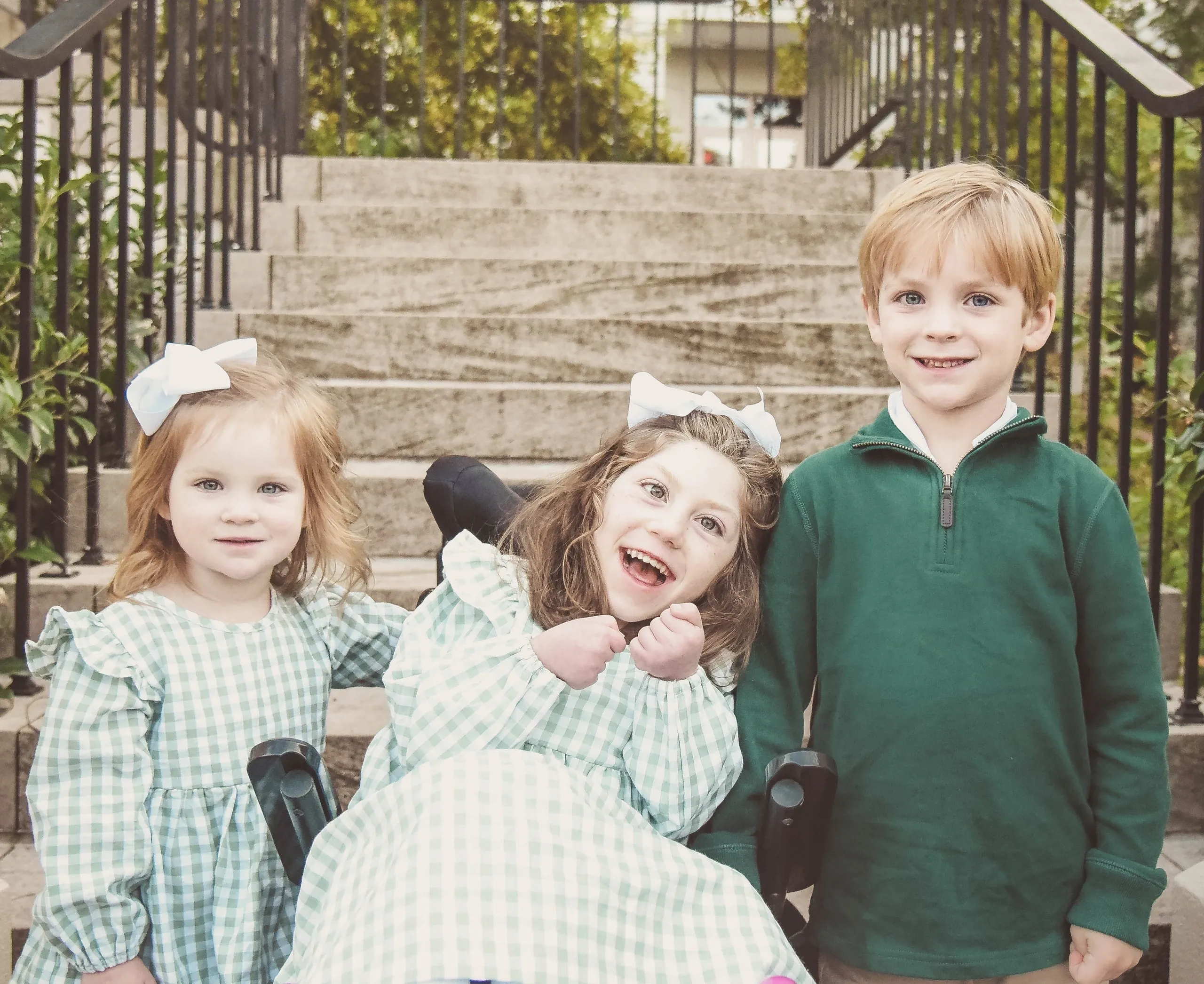 Three smiling children on outdoor steps, two girls in green gingham dresses and a boy in green, center girl in a wheelchair