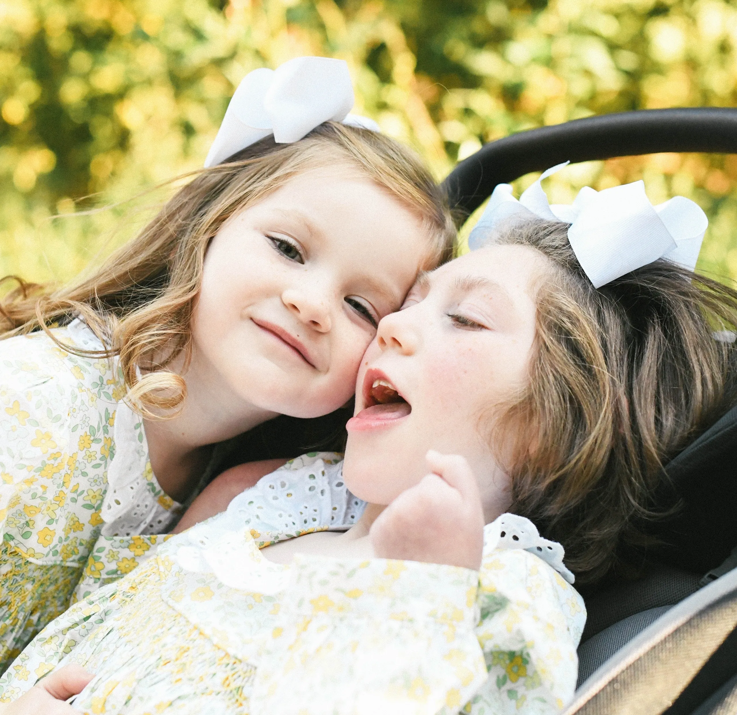 Close-up of two young girls with white bows cuddling in matching yellow floral dresses outdoors