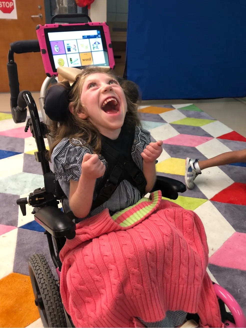 A child laughing in a classroom, a tablet with icons mounted behind, pink blanket over lap on a colorful rug.