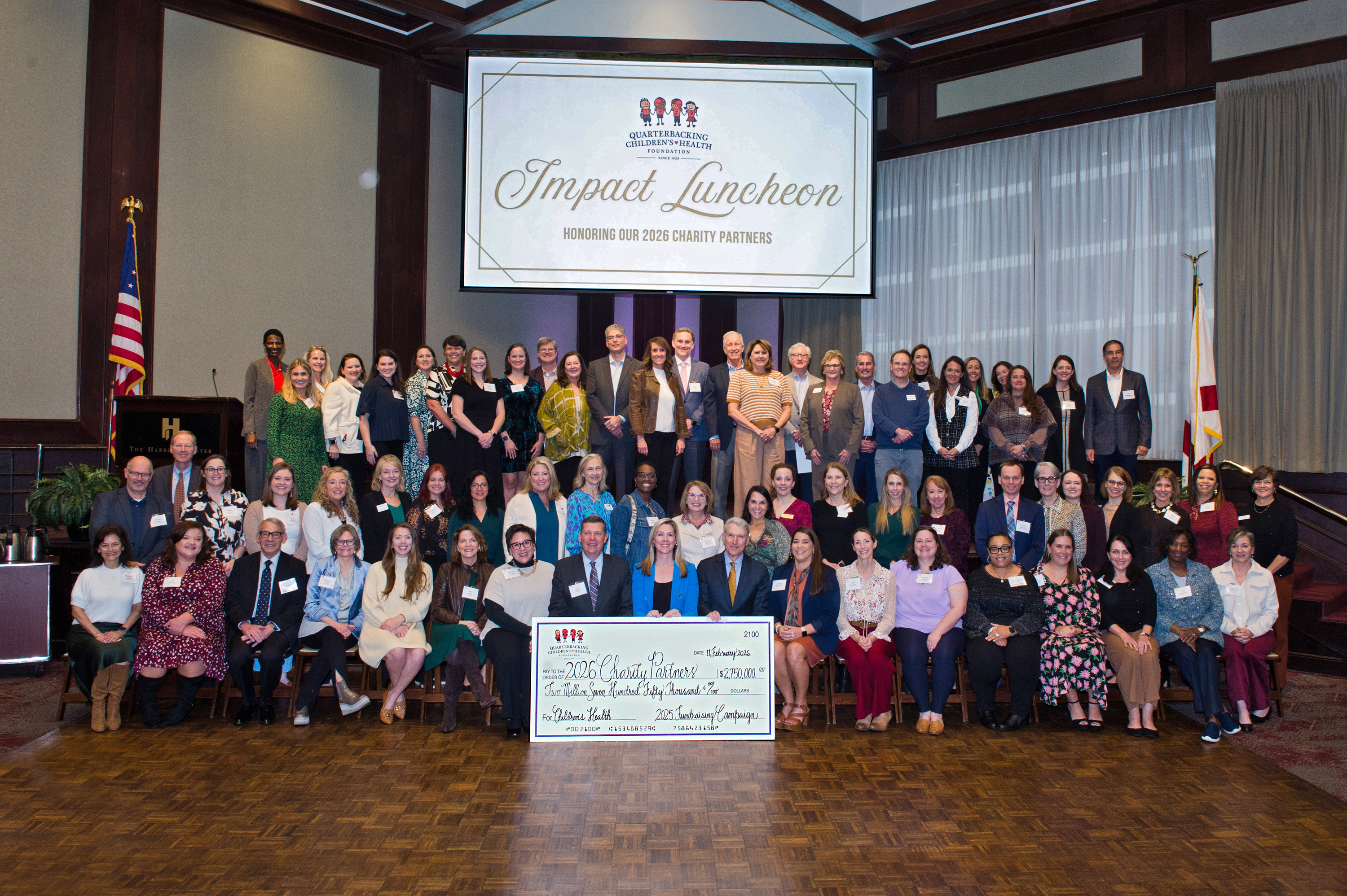 Large group at Impact Luncheon charity event posing with oversized check beneath sign honoring 2026 charity partners.