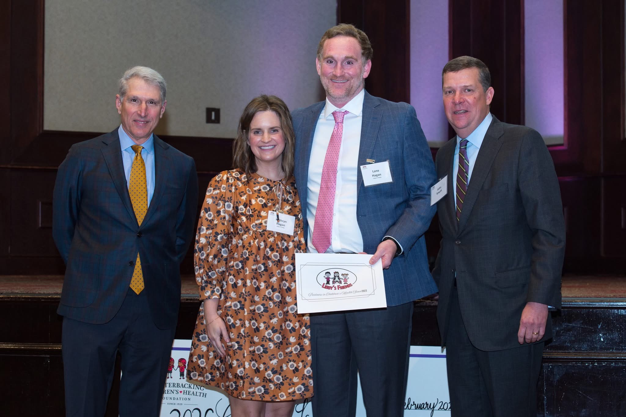 Four people at an award ceremony; a man holds a certificate while posing with colleagues in business attire on stage.