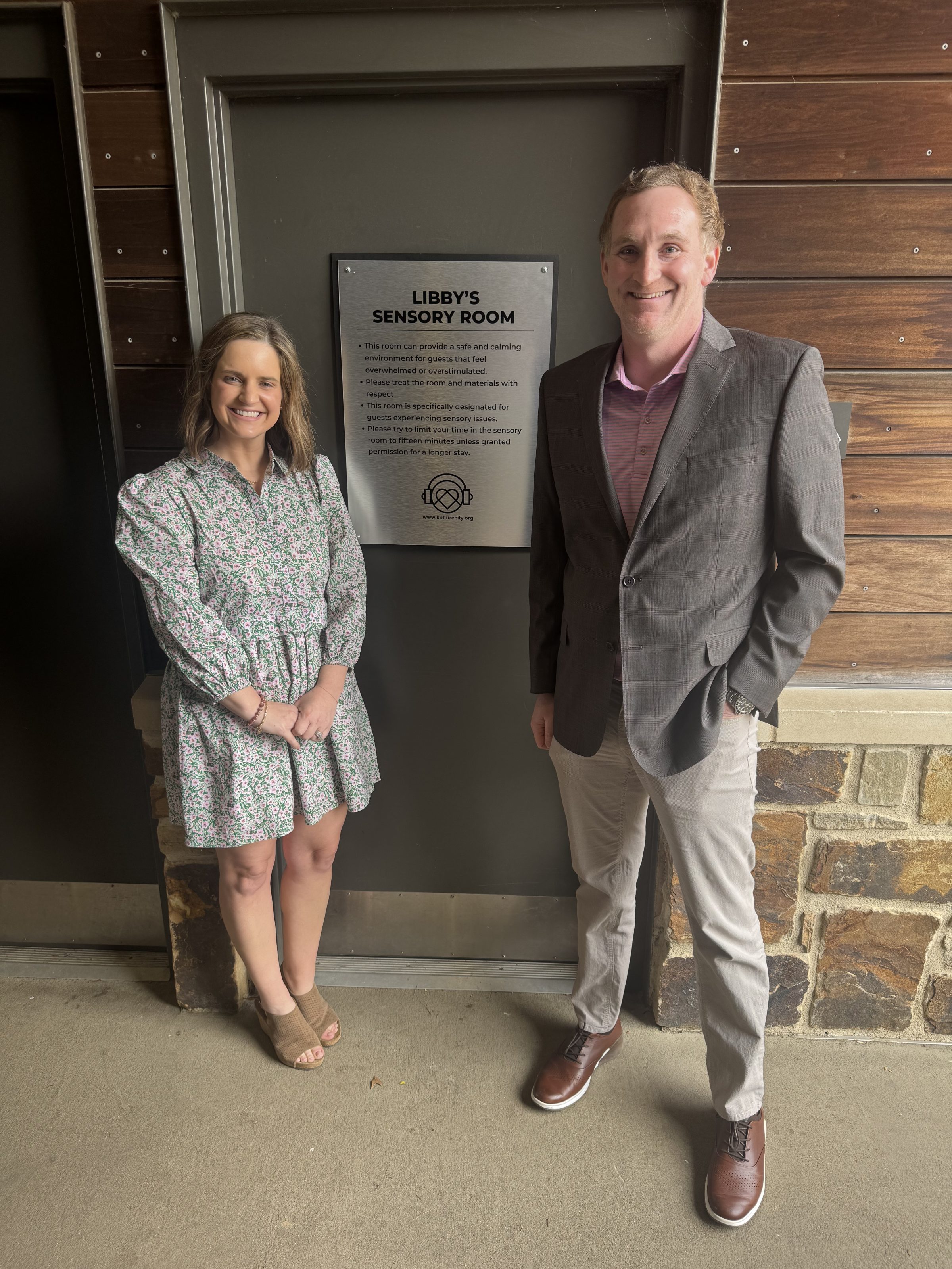 Two people, a woman in a floral dress and a man in a blazer, by a door labeled Libby's Sensory Room.