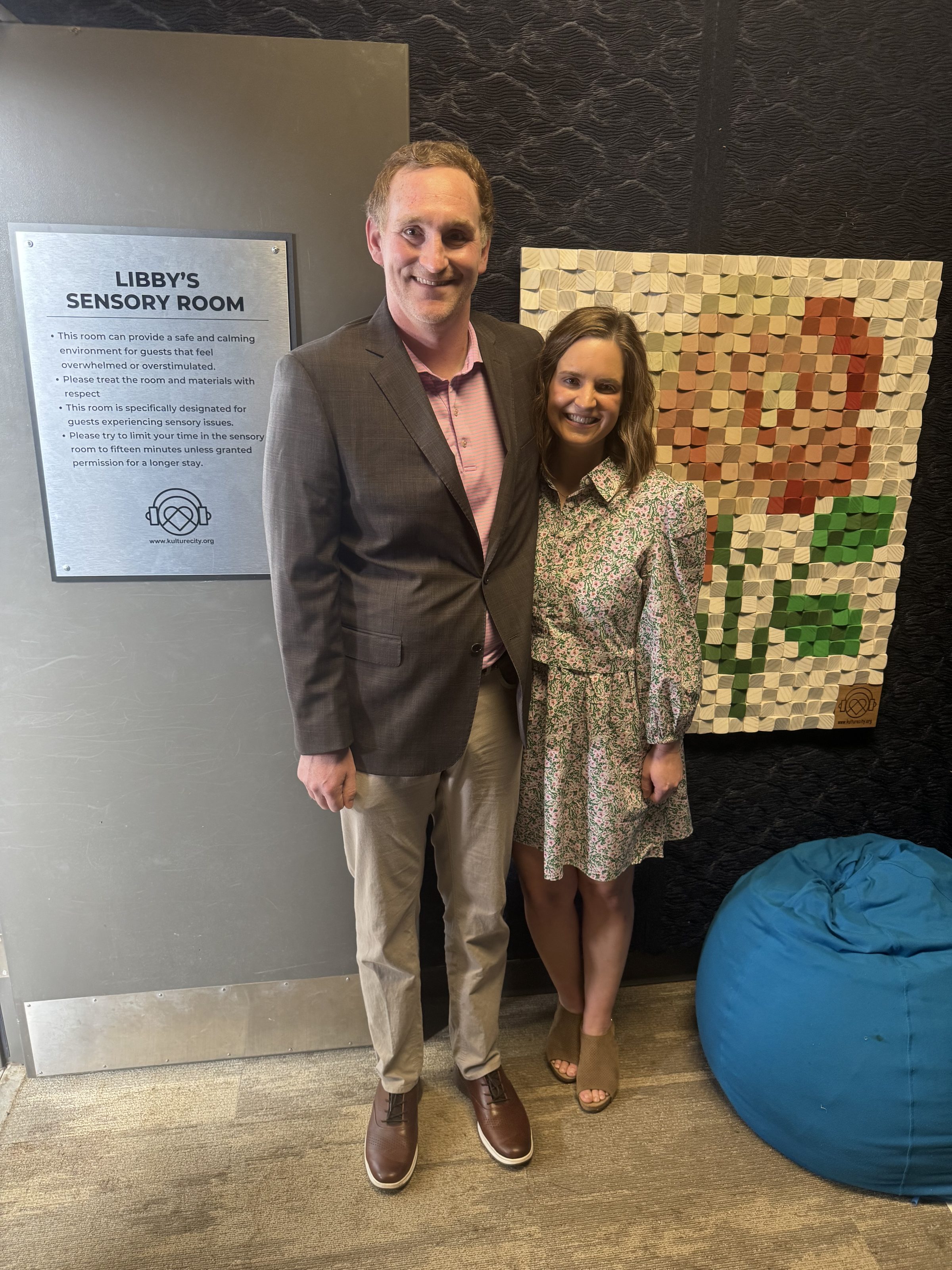 A man and a woman smiling by a 'Libby’s Sensory Room' sign, mosaic rose artwork, and a blue beanbag chair.