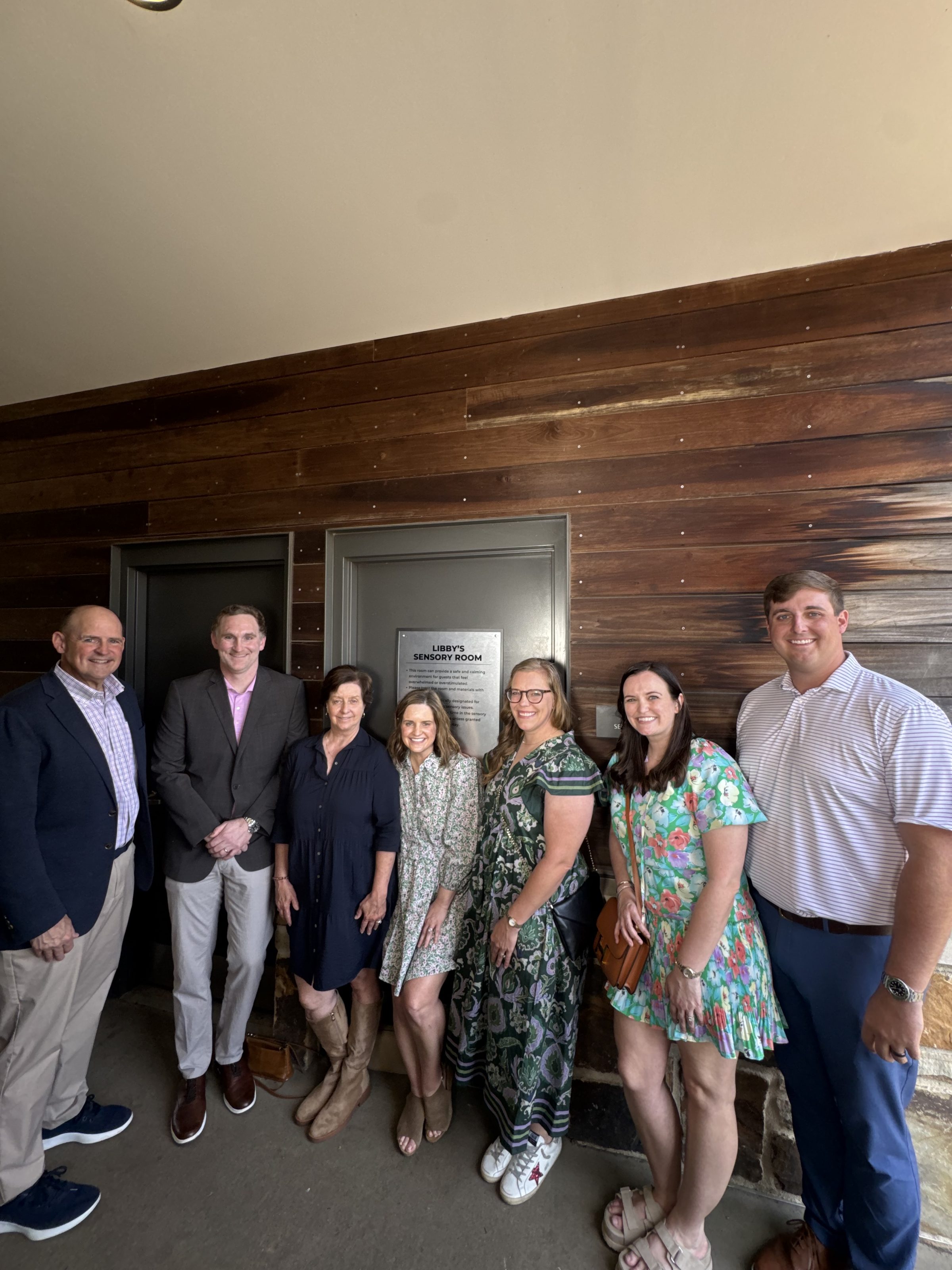 Group photo of seven adults smiling by a wooden wall outside a door labeled 'Libby's Sensory Room'.