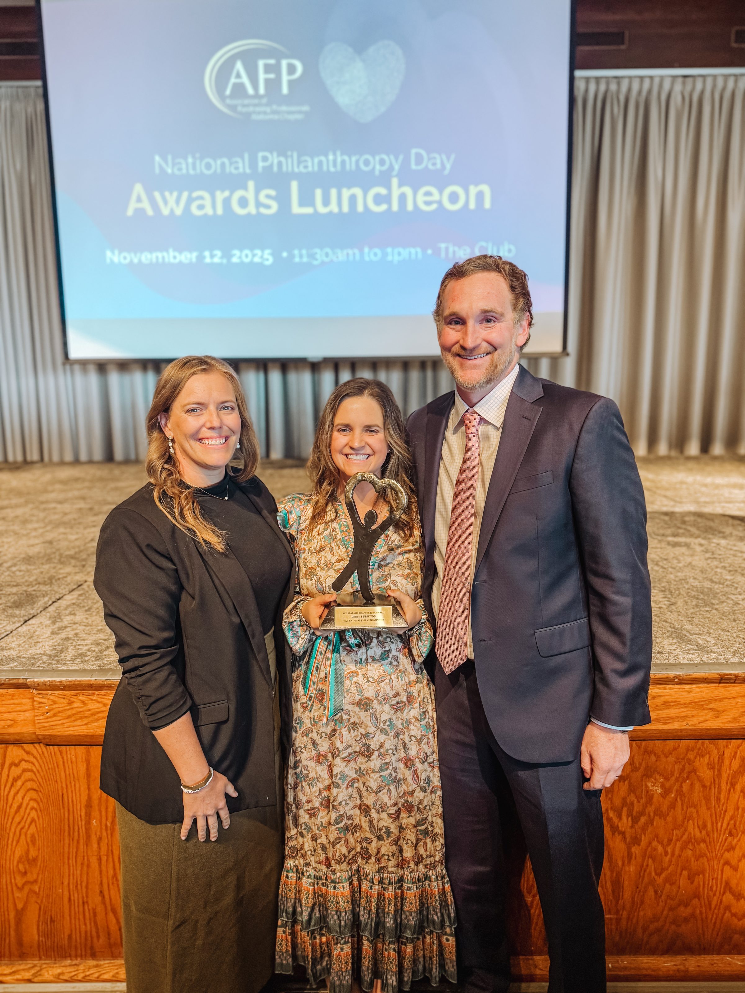 Three people smiling with an award at National Philanthropy Day Awards Luncheon, AFP event stage in background